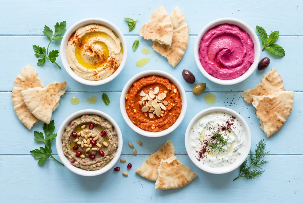 Cinco cuencos blancos con hummus, labni de betabel, feta con pimiento rojo, babaganoush y tzatziki sobre una mesa azul claro